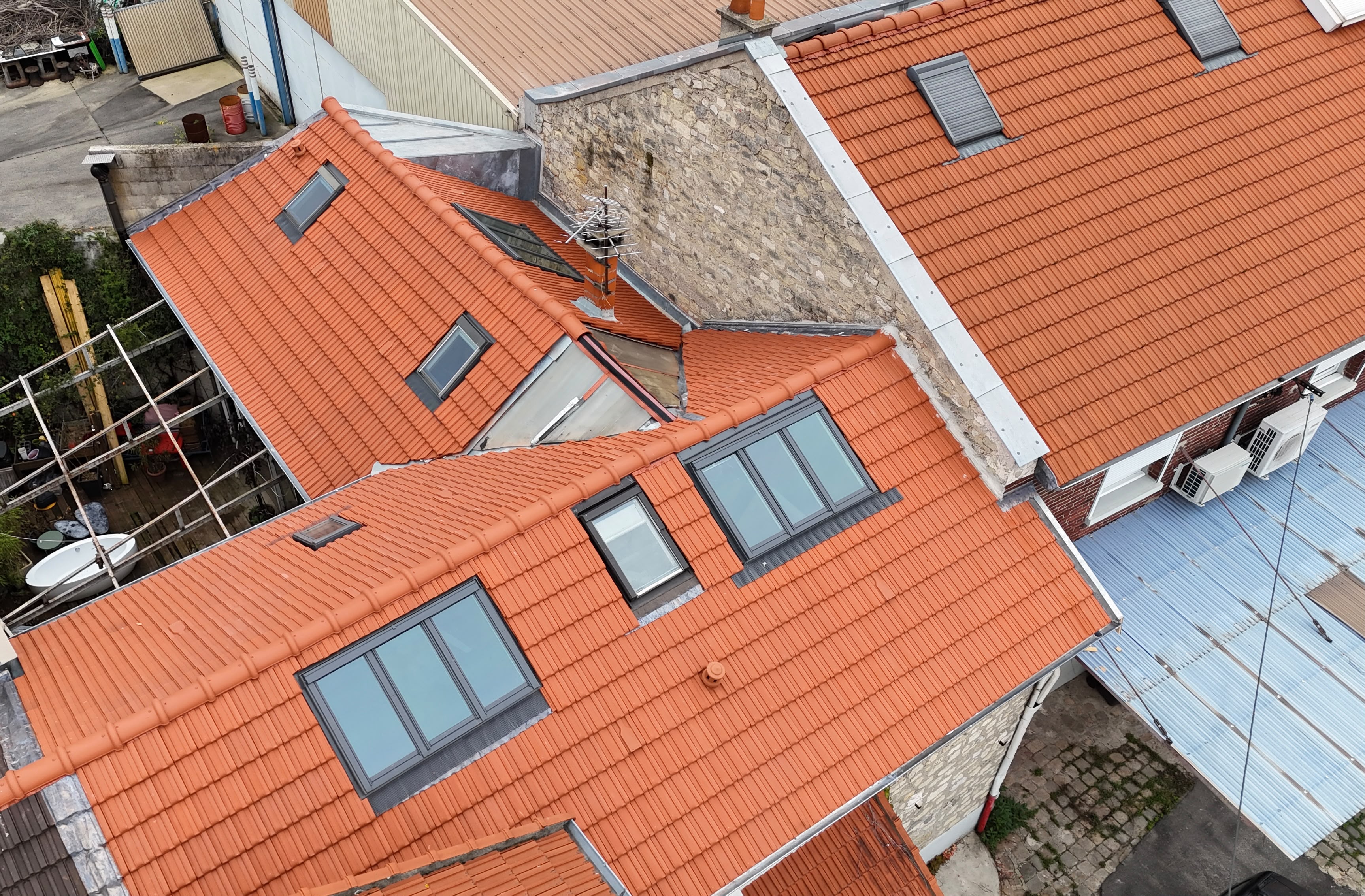 Toitures en tuiles orangées avec des fenêtres de toit et des raccords métalliques contre un mur en pierre à Bagnolet