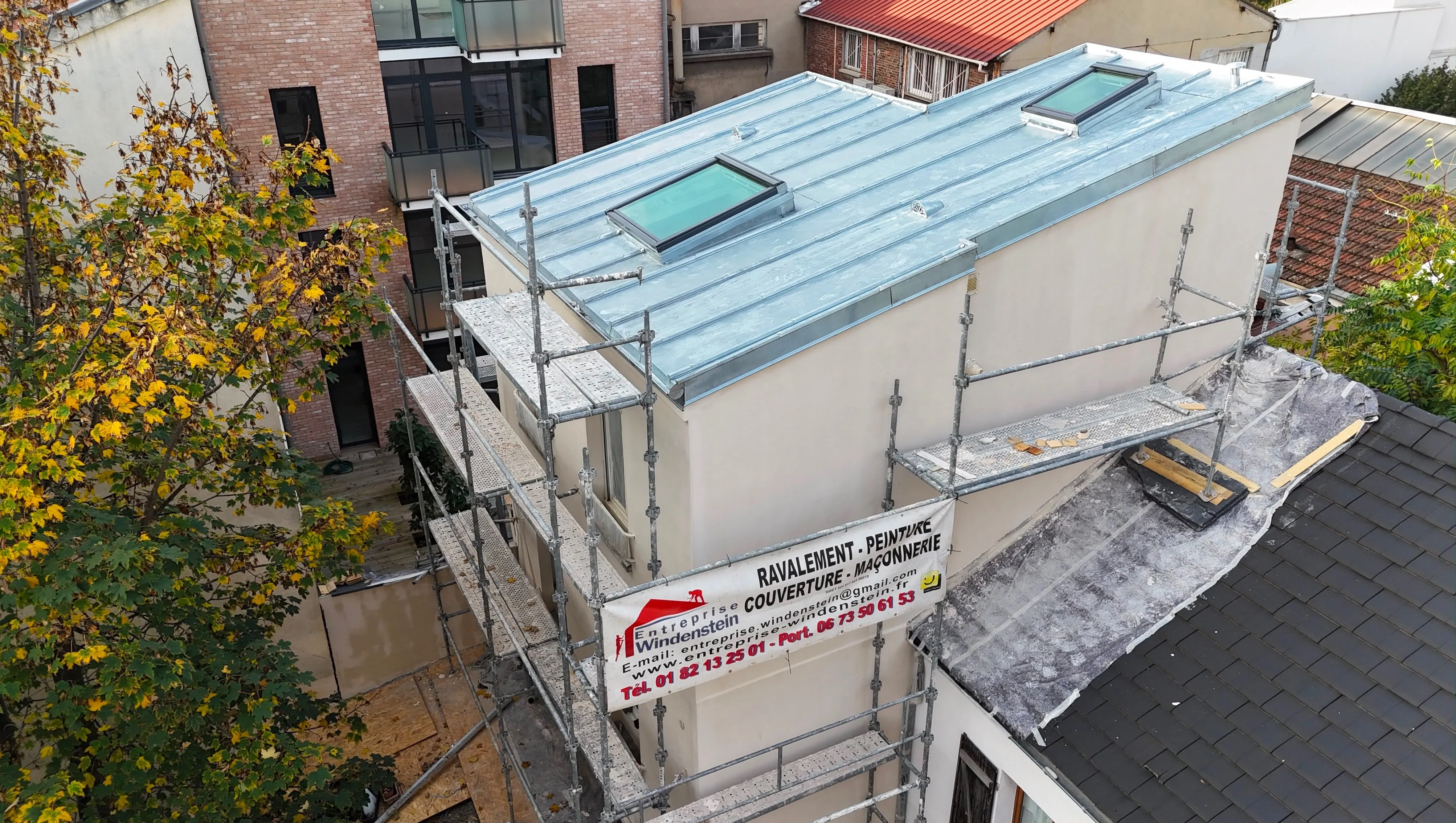 Toiture grise à reliefs parallèles avec deux fenêtres de toit et un échafaudage devant une façade claire